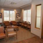 Comfortable patient waiting area with leather chairs, large windows, warm wood tones, and framed artwork on walls.