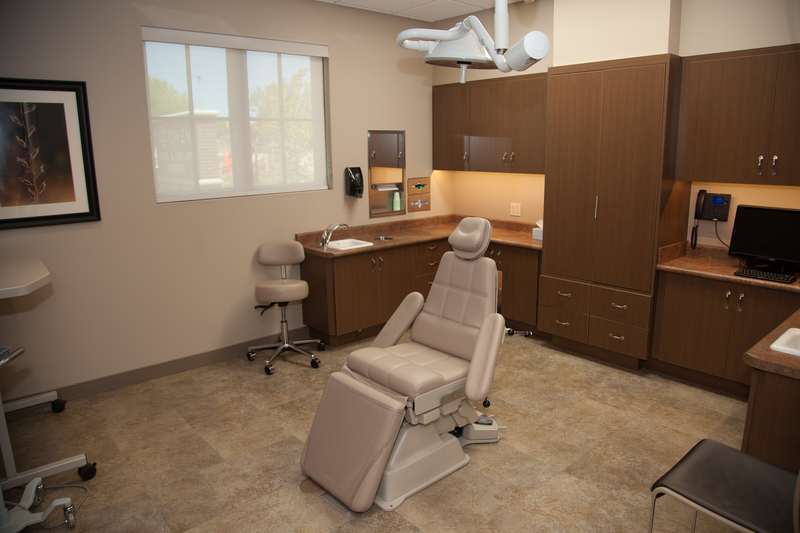 Spacious treatment room featuring white dental chair, overhead delivery system, wooden cabinetry, and large windows with natural lighting.