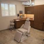 Spacious treatment room featuring white dental chair, overhead delivery system, wooden cabinetry, and large windows with natural lighting.