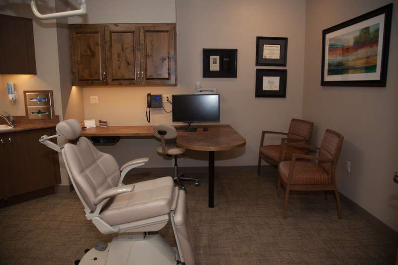 Modern dental treatment room equipped with white patient chair, computer workstation, cabinetry, and framed artwork.