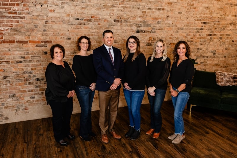 Professional team photo of seven staff members from Exceptional Smiles Family Dentistry standing together in front of an exposed brick wall, featuring one male team member in a blazer and tie at center with six female team members in dark tops and jeans, all smiling in a modern office waiting area.
